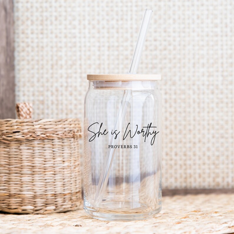 May include: A clear glass jar with a wooden lid and a clear straw. The jar has the words "She is Worthy" and "Proverbs 31" in black script. A woven basket is in the background.