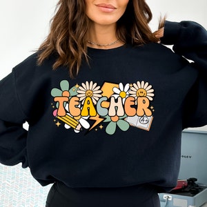 May include: Black sweatshirt with a graphic design of the word "TEACHER" in colorful, retro-style letters surrounded by flowers, stars, a pencil, a lightning bolt, and a book with an A+ grade.