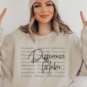 May include: A beige crewneck sweatshirt featuring black text that lists various positive adjectives including Purposeful, Impactful, Authentic, Influential, Selfless, Inspiring, Motivated, Passionate, Resilient, Driven, Empowering, Caring, Bold, Visionary, Dedicated, Courageous, Determined, Strategic, Proactive, Innovative, Transformative, Kind, Supportive, Encouraging, Guiding, Grateful, Patient, Understanding, with the words Difference Maker written in a larger black script font.