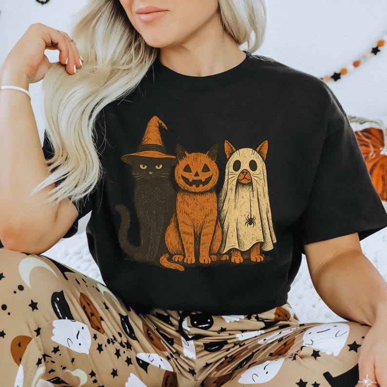 K&ouml;nnte beinhalten: Schwarzes T-Shirt mit einem Halloween-Katzen-Design. Die Grafik zeigt eine schwarze Katze mit Hexenhut, eine orangefarbene Katze mit K&uuml;rbisgesicht und eine Katze im Geisterkost&uuml;m. Die Farben sind Orange, Schwarz und Beige. Ein Artikel zum Thema Halloween.