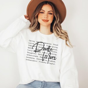 May include: White sweatshirt with black text that reads "Caring. Active. Generous. Proud. Loving. Attentive. Treat-giving. Indulgent. Devourous. Gentle. Protective. Responsible. Furry-obsesed. Social. Walking. Thoughtful. Nurturing. Spoiling. Affectionate. Supportive. Happy. Outdoorsy. Encouraging. Cuddles. Enthusiastic. Friendly. Playful." with the words "Doodle Mom" in a larger font.