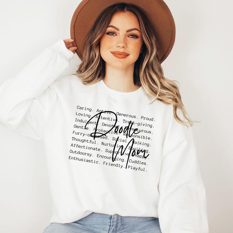May include: White sweatshirt with black text that reads "Caring. Active. Generous. Proud. Loving. Attentive. Treat-giving. Indulgent. Devourous. Gentle. Protective. Responsible. Furry-obsesed. Social. Walking. Thoughtful. Nurturing. Spoiling. Affectionate. Supportive. Happy. Outdoorsy. Encouraging. Cuddles. Enthusiastic. Friendly. Playful." with the words "Doodle Mom" in a larger font.