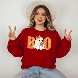 以下が含まれることがあります： オレンジ色の文字で「BOO」と書いてあり、クモの巣模様の赤いスウェットシャツ。ゴーストがカボチャを持っています。