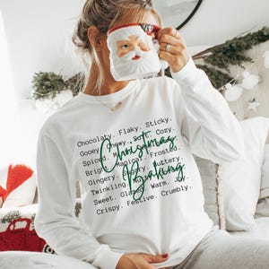 Pu&ograve; includere: Una maglietta bianca a maniche lunghe con testo nero che elenca parole come Chocolaty, Flaky, Sticky, Gooey, Chewy, Soft, Cozy, Spiced, Magical, Frosted, Bright, Gingery, Sugary, Buttery, Twinkling, Nutty, Warm, Joy, Sweet, Glazed, Crispy, Festive, Crumbly, con la scritta in corsivo verde Christmas Baking sovrapposta.