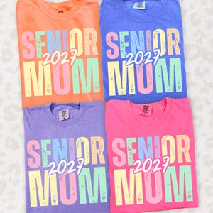 Puede incluir: Cuatro camisetas coloridas en naranja, azul, morado y rosa. Cada camiseta presenta el texto "SENIOR 2027 MUM" en una fuente multicolor desgastada. Las camisetas son probablemente de algod&oacute;n y de estilo casual.