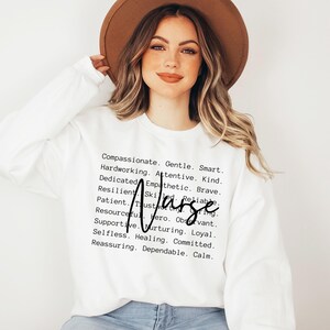 K&ouml;nnte beinhalten: Ein wei&szlig;er Pullover mit dem Wort "Krankenschwester" in fetten schwarzen Buchstaben und einer Liste positiver Attribute, die den Beruf beschreiben. Die Attribute beinhalten W&ouml;rter wie mitf&uuml;hlend, sanft, klug, flei&szlig;ig, aufmerksam, freundlich, engagiert, empathisch, mutig, belastbar, geschickt, zuverl&auml;ssig, geduldig, vertrauensw&uuml;rdig, aufmerksam, einfallsreich, Held, loyal, selbstlos, heilend, engagiert, beruhigend, zuverl&auml;ssig und ruhig.
