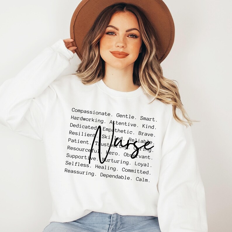 K&ouml;nnte beinhalten: Ein wei&szlig;er Pullover mit dem Wort "Krankenschwester" in fetten schwarzen Buchstaben und einer Liste positiver Attribute, die den Beruf beschreiben. Die Attribute beinhalten W&ouml;rter wie mitf&uuml;hlend, sanft, klug, flei&szlig;ig, aufmerksam, freundlich, engagiert, empathisch, mutig, belastbar, geschickt, zuverl&auml;ssig, geduldig, vertrauensw&uuml;rdig, aufmerksam, einfallsreich, Held, loyal, selbstlos, heilend, engagiert, beruhigend, zuverl&auml;ssig und ruhig.
