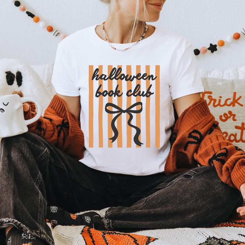 May include: White t-shirt with the text "halloween book club" in black script, set against orange and white vertical stripes. A black bow graphic is below the text. The shirt is part of a fall-themed collection.