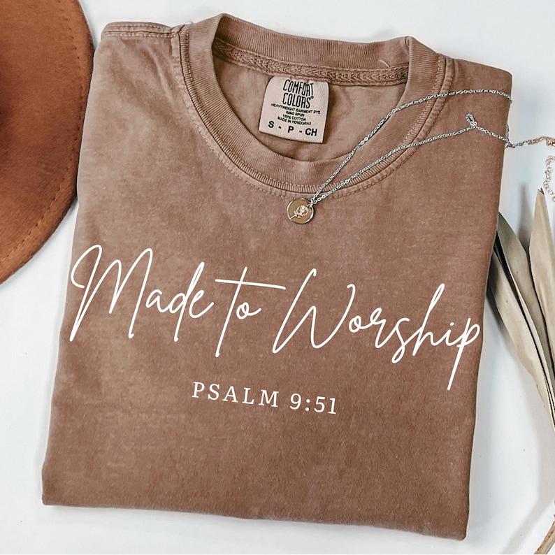 May include: A brown Comfort Colors t-shirt with the words "Made to Worship" in white script. The text "Psalm 9:51" is below the script. A silver necklace with a small pendant rests on the shirt.