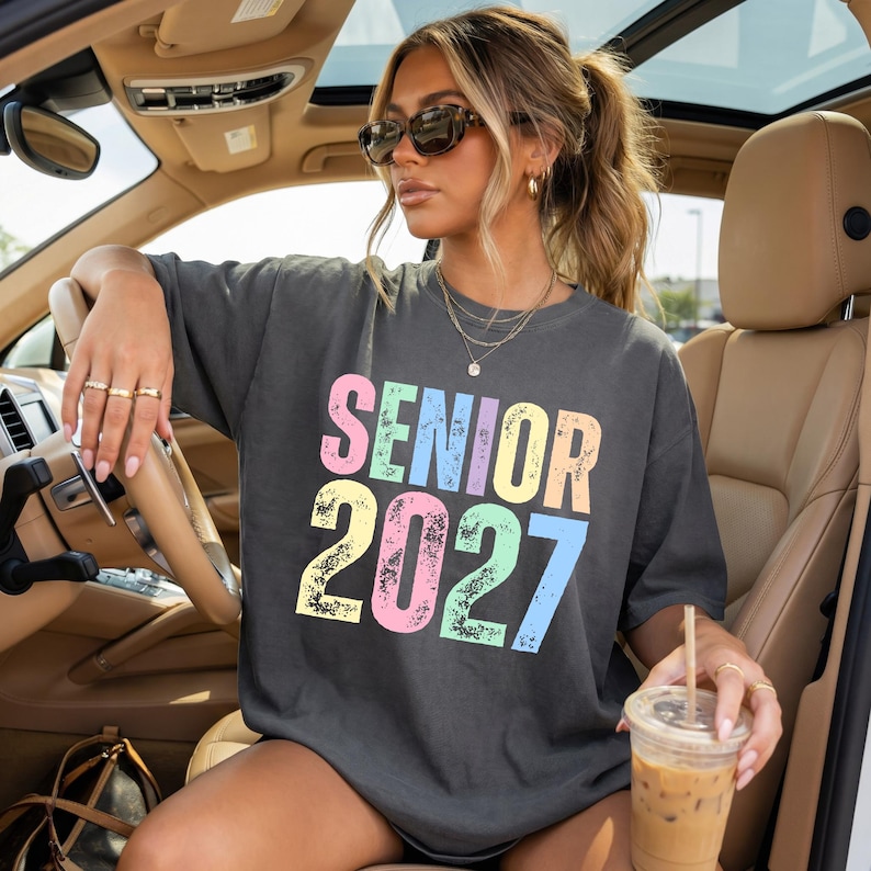 May include: A charcoal gray short-sleeve t-shirt featuring the text SENIOR 2027 printed in a distressed, multi-colored font. The letters and numbers appear in pastel shades of pink, blue, yellow, and green against the dark fabric background.