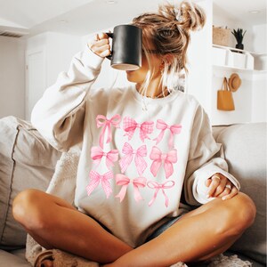 Puede incluir: Sudadera beige con ilustraciones de lazos rosas. Los lazos var&iacute;an en estilo, algunos con estampado de cuadros. La sudadera tiene mangas largas y cuello redondo. Una taza negra se sostiene frente a la cara.