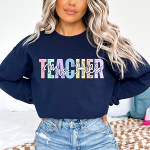 K&ouml;nnte beinhalten: Marineblaues Sweatshirt mit dem Wort "TEACHER" in Pastellfarben und dem Namen "Mrs. Hill" in Schreibschrift. Das Sweatshirt hat einen Rundhalsausschnitt und ist aus einem weichen Material gefertigt.