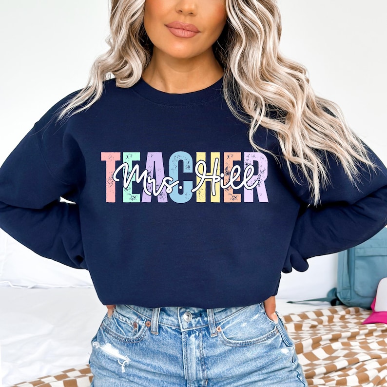K&ouml;nnte beinhalten: Marineblaues Sweatshirt mit dem Wort "TEACHER" in Pastellfarben und dem Namen "Mrs. Hill" in Schreibschrift. Das Sweatshirt hat einen Rundhalsausschnitt und ist aus einem weichen Material gefertigt.