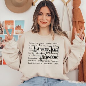 May include: A beige sweatshirt with the text "Homestead Mama" in black lettering. The text is surrounded by a list of words describing the qualities of a homesteader, such as "animal caretaker", "handy", "rural", "strong-willed", "gardener", "DIYer", "resilient", "resourceful", "patient", "homemade", "earth", "sustainable", "nurturing", "self-sufficient", "preserver", "holistic", "creative", "independent", "forager", "back-to-basics", "hardwork", and "grow".