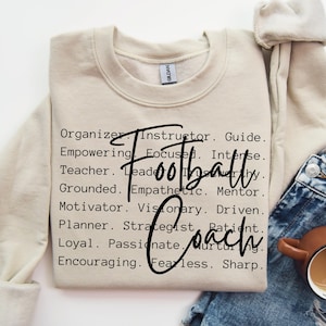 May include: A beige sweatshirt with the words "Football Coach" in black text. The words are surrounded by a list of positive attributes, such as "Organizer, Instructor, Guide, Empowering, Focused, Intense, Teacher, Leader, Grounded, Empathetic, Mentor, Motivator, Visionary, Driven, Planner, Strategist, Patient, Loyal, Passionate, Nurturing, Encouraging, Fearless, Sharp."