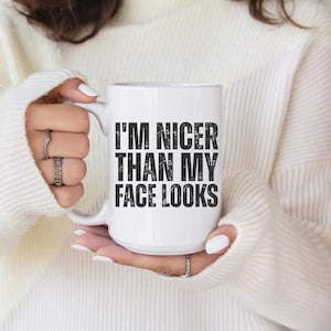 Pu&ograve; includere: Tazza in ceramica bianca con la scritta "I'M NICER THAN MY FACE LOOKS" in lettere nere. La tazza &egrave; tenuta da una persona che indossa un maglione bianco. La mano della persona &egrave; visibile, con diversi anelli alle dita.
