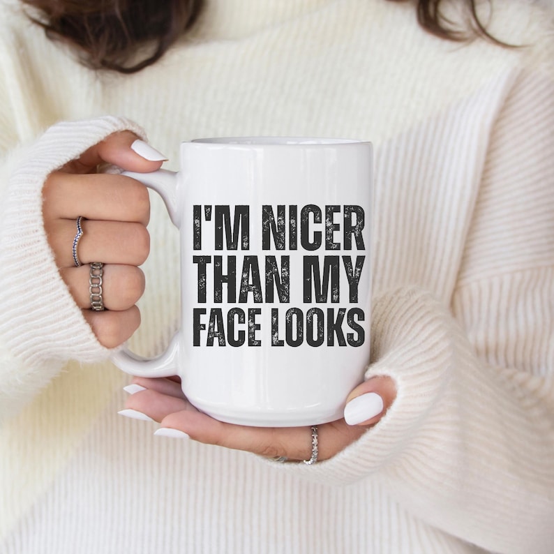 Pu&ograve; includere: Tazza in ceramica bianca con la scritta "I'M NICER THAN MY FACE LOOKS" in lettere nere. La tazza &egrave; tenuta da una persona che indossa un maglione bianco. La mano della persona &egrave; visibile, con diversi anelli alle dita.