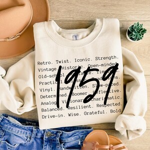 May include: Cream-colored sweatshirt with the year "1959" in large black numbers. The shirt features a list of words like "Retro," "Vintage," and "Iconic" in a black font. The sweatshirt is paired with blue jeans and a tan hat.
