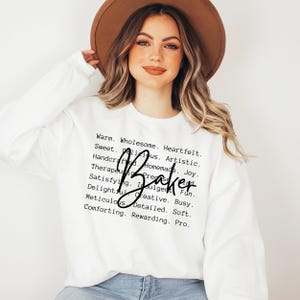 May include: White sweatshirt with the text "Warm. Wholesome. Heartfelt. Sweet. Delicious. Artistic. Handcrafted. Homemade. Joy. Therapeutic. Present. Fun. Satisfying. Indulgent. Busy. Delightful. Creative. Soft. Meticulous. Detailed. Pro." and the word "Baker" written in cursive.