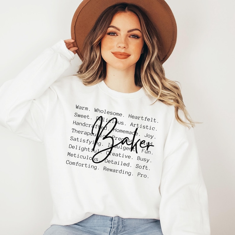 May include: White sweatshirt with the text "Warm. Wholesome. Heartfelt. Sweet. Delicious. Artistic. Handcrafted. Homemade. Joy. Therapeutic. Present. Fun. Satisfying. Indulgent. Busy. Delightful. Creative. Soft. Meticulous. Detailed. Pro." and the word "Baker" written in cursive.