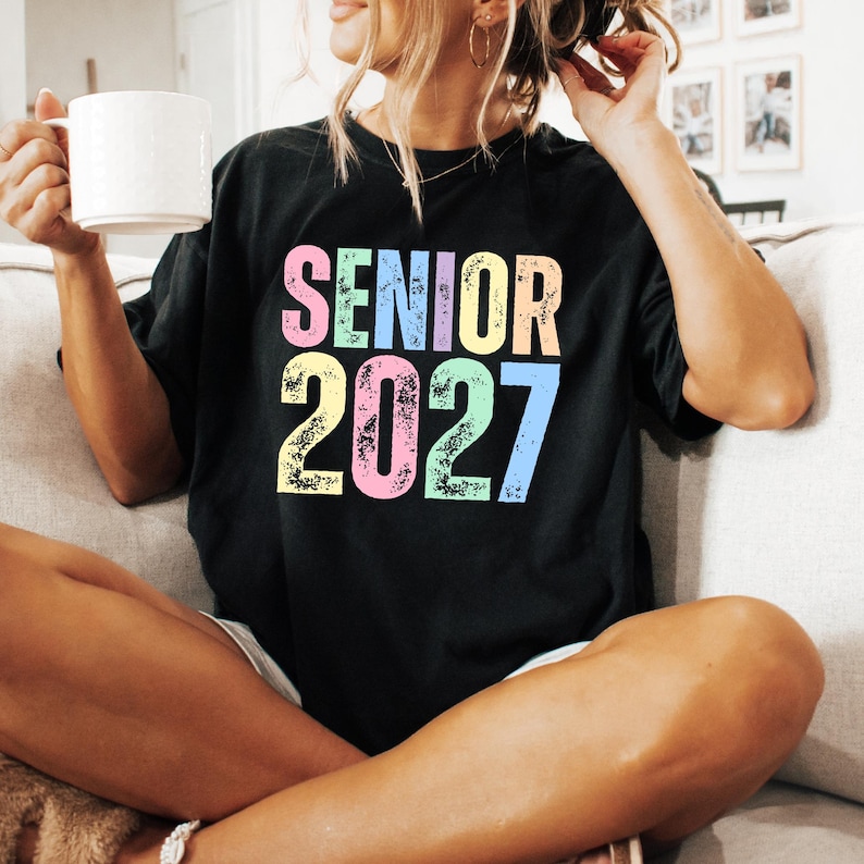 May include: A black short-sleeved t-shirt featuring the text SENIOR 2027 printed in a distressed, multi-colored font. The letters and numbers are colored in shades of pink, teal, yellow, and blue, creating a bold graphic design on the chest.