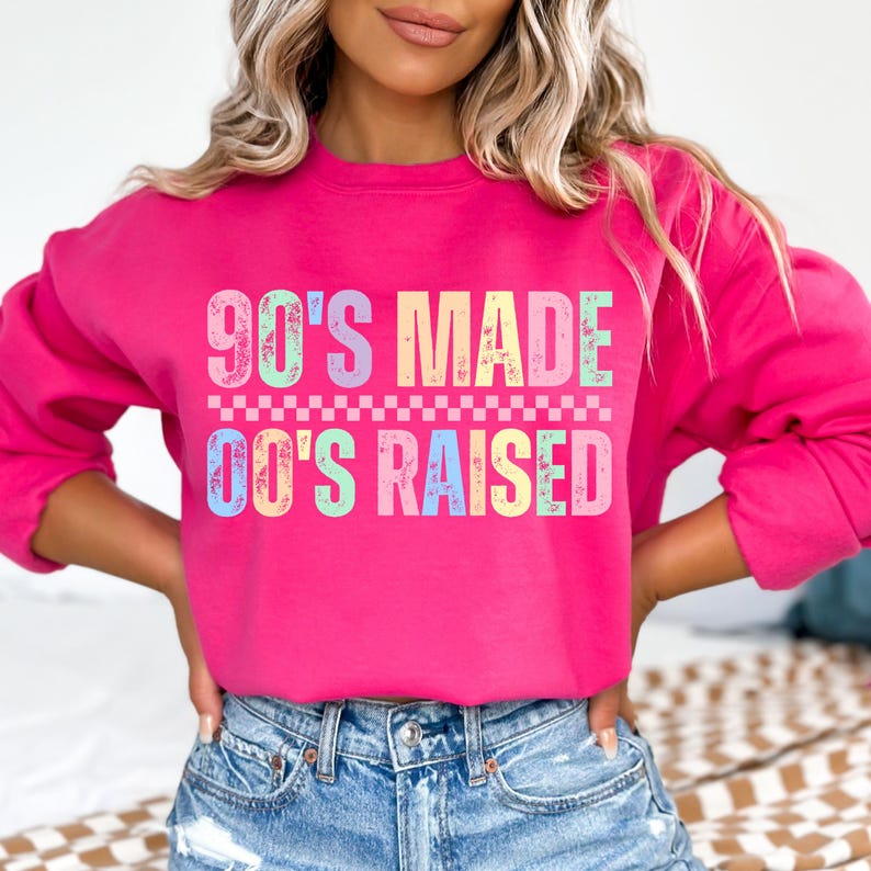 May include: A bright pink sweatshirt with the text "90'S MADE 00'S RAISED" in pastel colors. The text is above a checkered pattern. The sweatshirt is worn with blue denim shorts.