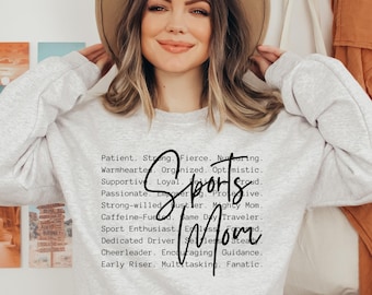 May include: A light gray crewneck sweatshirt featuring black text that lists various traits including Patient, Strong, Fierce, Nursing, Warmhearted, Organized, Optimistic, Supportive, Loyal, Proud, Passionate, Empowering, Protective, Strong-willed, Hustler, Mighty Mom, Caffeine-Fueled, Same Day Traveler, Sport Enthusiast, Endless, Focused, Dedicated Driver, Selfless, Steady, Cheerleader, Encouraging, Guidance, Early Riser, Multitasking, Fanatic, with the words Sports Mom written in a large black script font.