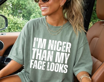 May include: A sage green short-sleeved t-shirt featuring the white distressed text I'M NICER THAN MY FACE LOOKS printed on the front. The shirt has a crew neckline and a relaxed fit, showcasing a casual style with bold, readable lettering.