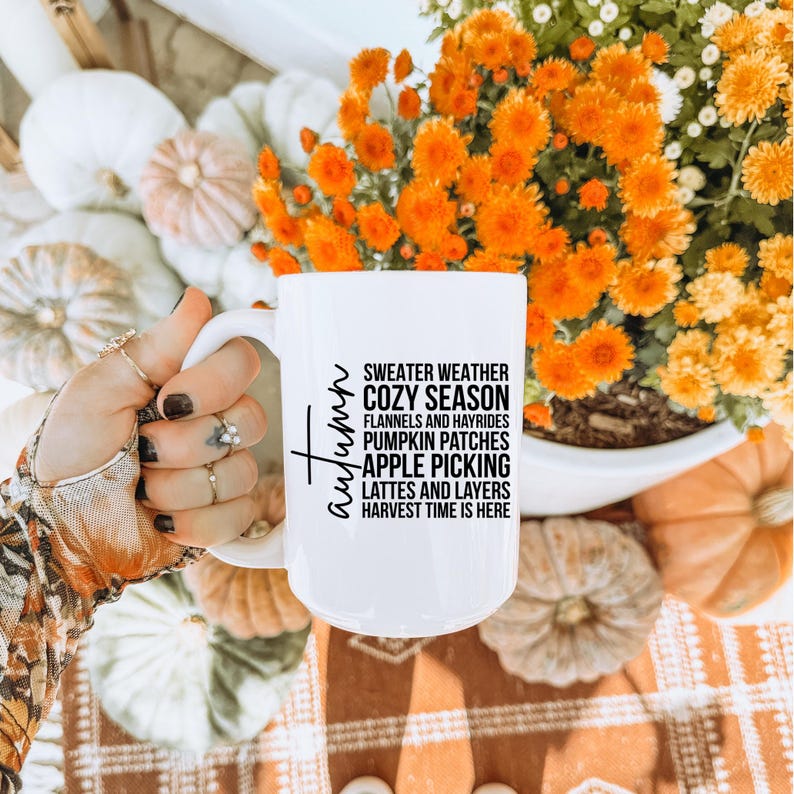 K&ouml;nnte beinhalten: Wei&szlig;e Keramiktasse mit dem Wort "Autumn" in Schrift und Text, der Herbstaktivit&auml;ten auflistet. Die Tasse wird in einer Hand mit Ringen gehalten, umgeben von K&uuml;rbissen und orangefarbenen Blumen.