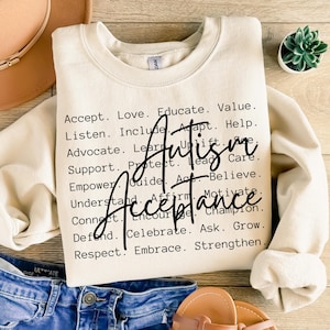 Pode incluir: Uma camisola de gola redonda cor de creme com texto preto que lista vários verbos positivos, incluindo Accept, Love, Educate, Value, Listen, Include, Adapt, Help, Advocate, Learn, Uplift, Support, Protect, Lead, Care, Empower, Guide, Act, Believe, Understand, Affirm, Motivate, Connect, Encourage, Champion, Defend, Celebrate, Ask, Grow, Respect, Embrace, Strengthen, com as palavras Autism Acceptance escritas numa fonte cursiva preta grande no centro.