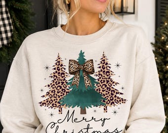 May include: A cream-colored crewneck sweatshirt featuring a graphic of three Christmas trees. The two outer trees have a leopard print pattern, while the center tree is solid green with a large leopard print bow. The text Merry Christmas is below.