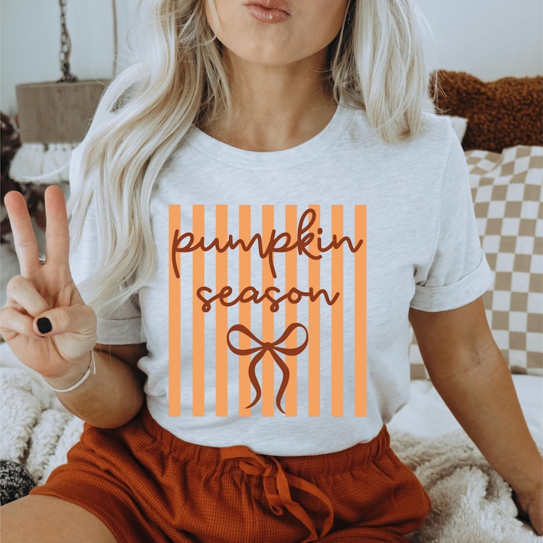 May include: A light gray t-shirt with the words "pumpkin season" in a brown script font, set against a background of orange vertical stripes. A brown bow graphic is below the text. The shirt is paired with orange shorts.