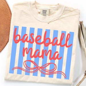 May include: Cream-colored t-shirt with the words "baseball mama" in red script over blue vertical stripes. A red bow is at the bottom of the design. The shirt has a crew neck and short sleeves.