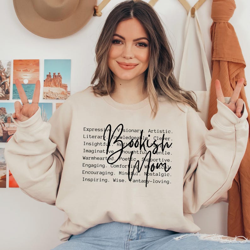 Puede incluir: Una mujer lleva una sudadera de color beige claro con el texto "Bookish Mom" en letras negras. El texto est&aacute; rodeado de una lista de palabras que describen las cualidades de una madre aficionada a la lectura, como "literaria,  culta,  perspicaz,  imaginativa,  reflexiva,  amable,  cari&ntilde;osa,  po&eacute;tica,  solidaria,  atractiva,  reconfortante,  alentadora,  consciente,  nost&aacute;lgica,  inspiradora,  sabia,  amante de la fantas&iacute;a."