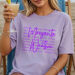 May include: A light purple t-shirt with the words "Margarita Weather" in a cursive font. The shirt has a list of words associated with the drink and the beach. A person is holding a cocktail.