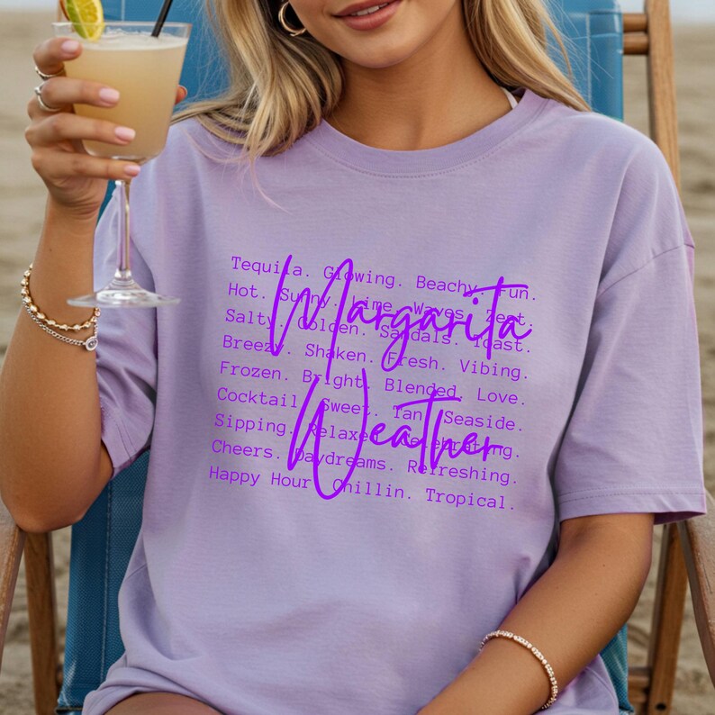 May include: A light purple t-shirt with the words "Margarita Weather" in a cursive font. The shirt has a list of words associated with the drink and the beach. A person is holding a cocktail.