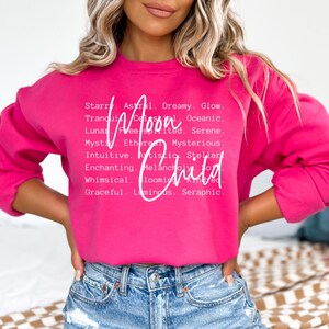 May include: A pink sweatshirt with the words "Moon Child" in white lettering. The words are surrounded by a list of adjectives describing the moon, such as "dreamy", "glow", "celestial", "oceanic", "free-spirited", "serene", "mysterious", "ethereal", "intuitive", "artistic", "stellar", "enchanting", "melancholy", "whimsical", "blooming", "kindred", "graceful", "luminous", and "seraphic".