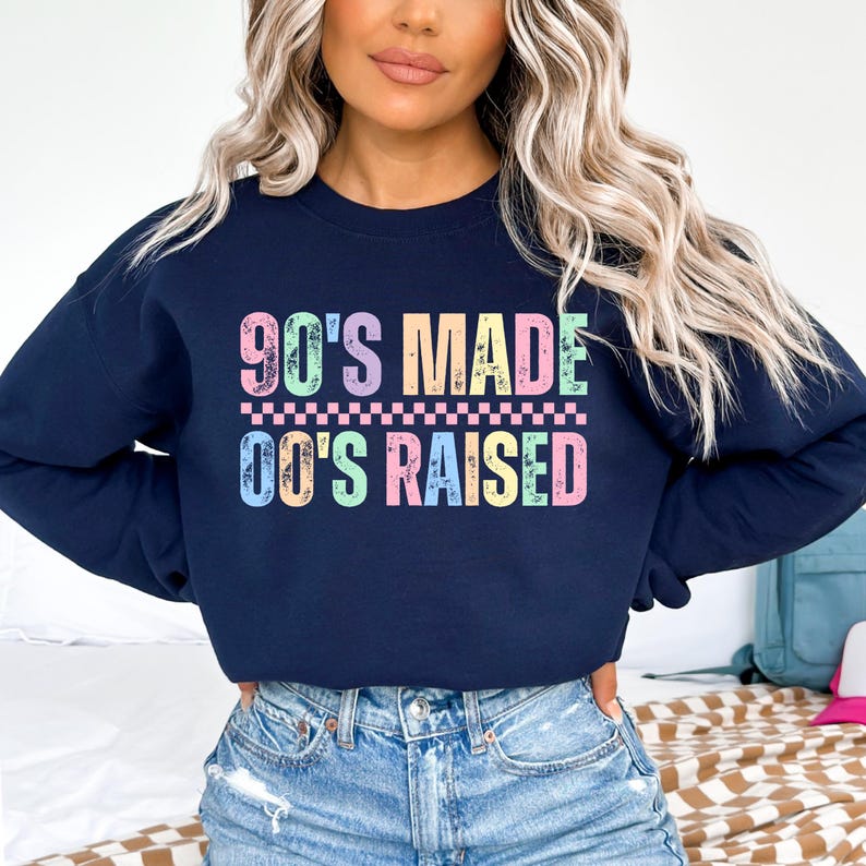 May include: Navy blue sweatshirt with the text "90'S MADE 00'S RAISED" in pastel colors. The text is in a retro font, with a checkered pattern between the two lines of text. The sweatshirt is a crew neck style.