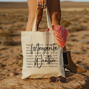 May include: A cream-colored tote bag with the word "Margarita" and "Weathering" printed on it. The bag is held by a person. A pink bandana and sunglasses are also visible.