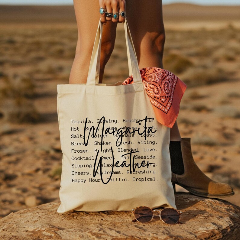 May include: A cream-colored tote bag with the word "Margarita" and "Weathering" printed on it. The bag is held by a person. A pink bandana and sunglasses are also visible.