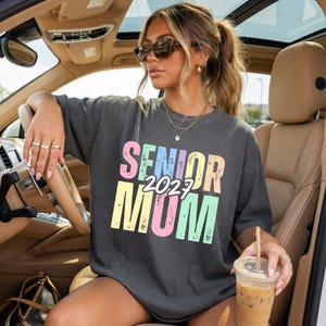 Puede incluir: Camiseta gris oscuro con la frase "SENIOR 2027 MOM" en colores pastel. La persona en el coche lleva gafas de sol. El texto es en negrita y f&aacute;cil de leer.