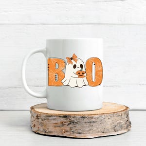 以下が含まれることがあります： 白いセラミック製のコーヒーカップ。かわいい幽霊がカボチャを持っており、オレンジ色の文字で「BOO」と書いてあり、クモの巣模様が描かれています。
