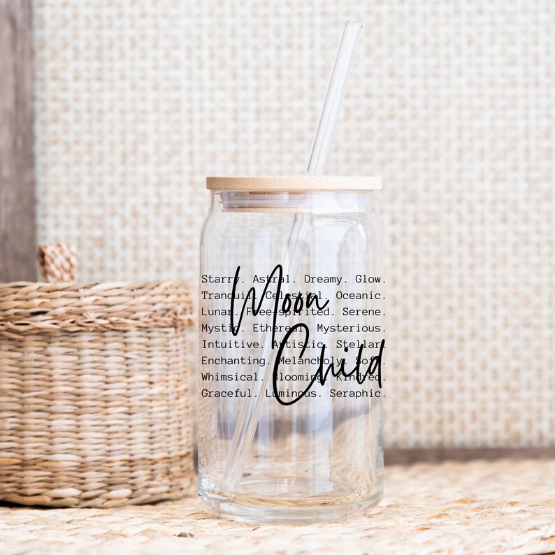May include: A clear glass jar with a wooden lid and a straw. The jar has a black and white design with the words "Moon Child" and a list of words describing the moon, such as "dreamy", "glow", "serene", and "mystic".