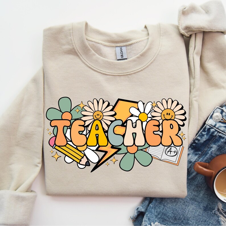 May include: A beige sweatshirt with a graphic design of the word "TEACHER" in colorful, retro-style letters surrounded by flowers, a lightning bolt, a pencil, and a notebook with an A+ grade.