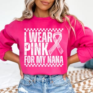 May include: A bright pink sweatshirt with the text "I WEAR PINK FOR MY NANA" in white, along with a white ribbon graphic. The sweatshirt has a crew neck and long sleeves. The text is framed by a white checkered pattern.