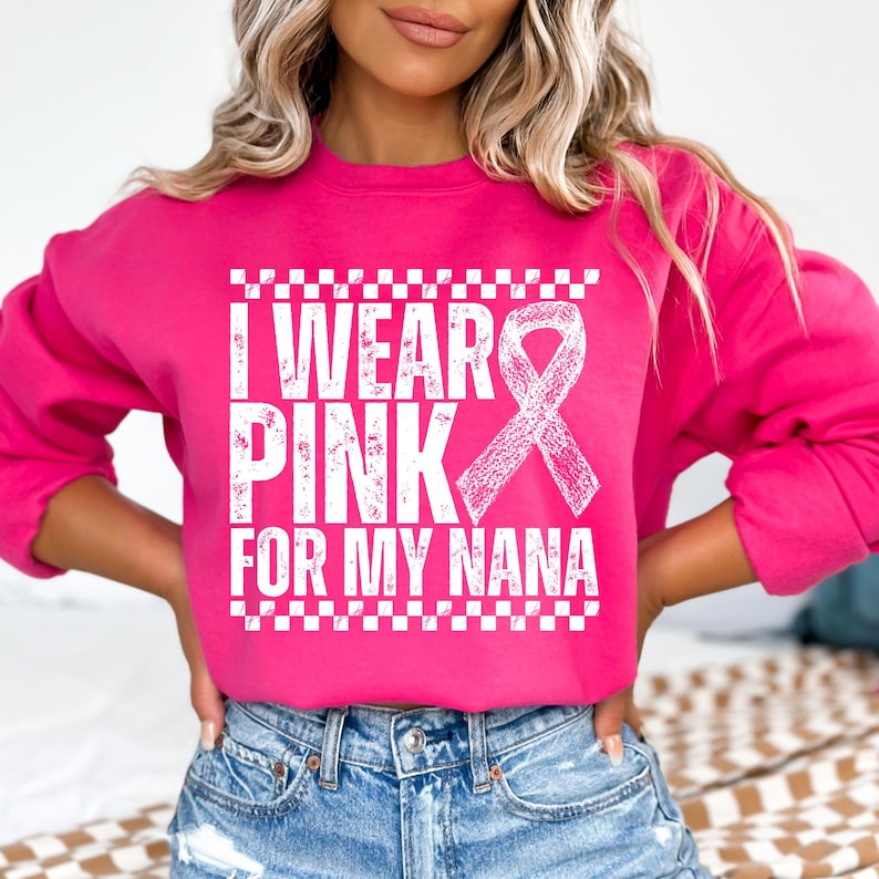 May include: A bright pink sweatshirt with the text "I WEAR PINK FOR MY NANA" in white, along with a white ribbon graphic. The sweatshirt has a crew neck and long sleeves. The text is framed by a white checkered pattern.