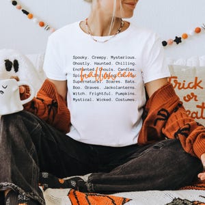 May include: White t-shirt with a Halloween-themed list of words in black font. The words include "Spooky," "Creepy," and "Mysterious." The shirt is worn with a burnt orange cardigan and dark jeans. The word "Halloween" is written in orange script.