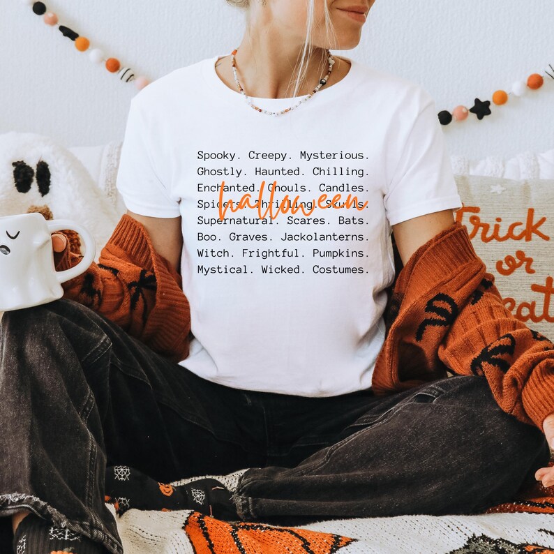May include: White t-shirt with a Halloween-themed list of words in black font. The words include "Spooky," "Creepy," and "Mysterious." The shirt is worn with a burnt orange cardigan and dark jeans. The word "Halloween" is written in orange script.