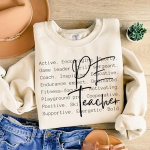 May include: Cream-colored sweatshirt with the words "PE Teacher" in a script font. The sweatshirt has a list of positive attributes such as "Active," "Encouraging," and "Sporty" printed in black.