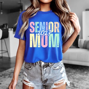 Puede incluir: Una camiseta azul real con las palabras "SENIOR 2023 MOM" en colores pastel. El texto est&aacute; centrado en la camiseta. La camiseta se lleva con pantalones cortos vaqueros.